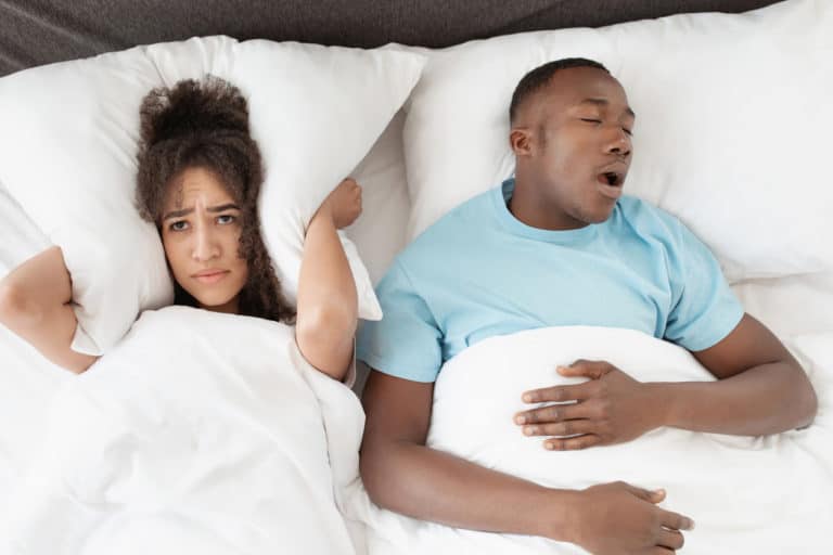 Woman covers her ears with a pillow while a man snores in bed beside her.