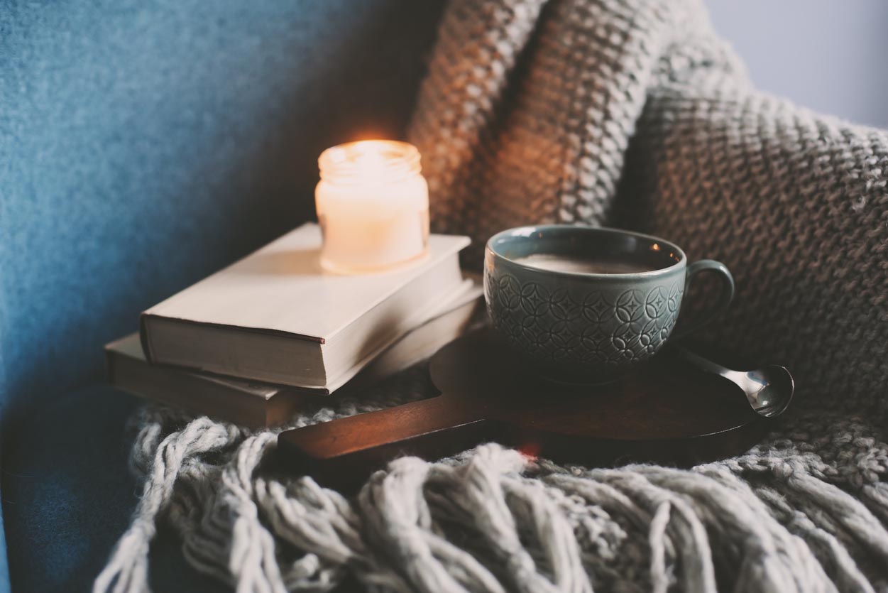 A cosy scene of a blanket, cup of tea, books and a candle on a comfy chair.